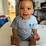 toddler, child, smiling, curly_hair, grey_tshirt, denim_shorts, bracelet, indoors, sitting, happy, face, eyes, mouth_open, skin, hand, footwear, soft_surface, background_blur, person, portrait