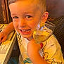 child, boy, smiling, glass, drink, table, menu, shirt, colorful_clothing, crab_print, duck_print, indoor, warm_lighting, face, happy, sitting, hair, hand, restaurant, wicker_chair