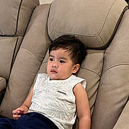 Kieran is registered to the contest to win money with this photo: child, toddler, portrait, sitting, recliner, chair, indoors, home, living_room, face, dark_hair, looking_away, calm, clothing, shirt, shorts, barefoot, furniture, relaxed, photography