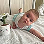baby, infant, bed, white_bedspread, plush_toy, flower_pot, blue_eyes, curious_expression, white_clothing, indoor, wooden_wardrobe, door, child, person, toddler, cute, soft_toy, smiling_toy, home, laying_down
