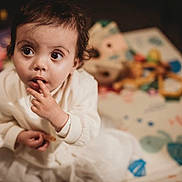 Maya joined the competition — help win amazing prizes! toddler, child, white_clothing, curious, indoor, play_mat, toy, blurred_background, person, face, hand, sitting, young_child, expression, cute, small_hands, short_hair, indoor_lighting, playtime, childhood