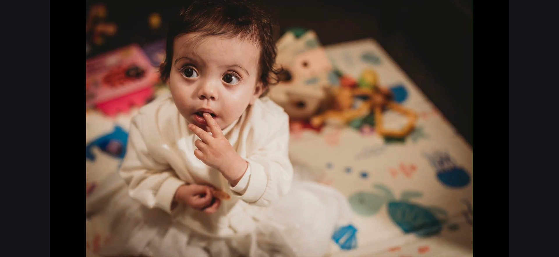 Maya joined the competition — help win amazing prizes! toddler, child, white_clothing, curious, indoor, play_mat, toy, blurred_background, person, face, hand, sitting, young_child, expression, cute, small_hands, short_hair, indoor_lighting, playtime, childhood