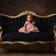 Ziyah is registered to the contest to win money with this photo: toddler, child, dress, pink_dress, sofa, vintage_sofa, black_sofa, gold_frame, bow, hair_accessory, portrait, indoor, studio, wood_floor, curious, seated, furniture, elegant, baby, young_child