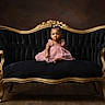 toddler, child, dress, pink_dress, sofa, vintage_sofa, black_sofa, gold_frame, bow, hair_accessory, portrait, indoor, studio, wood_floor, curious, seated, furniture, elegant, baby, young_child