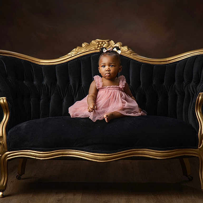 Ziyah is registered to the contest to win money with this photo: toddler, child, dress, pink_dress, sofa, vintage_sofa, black_sofa, gold_frame, bow, hair_accessory, portrait, indoor, studio, wood_floor, curious, seated, furniture, elegant, baby, young_child