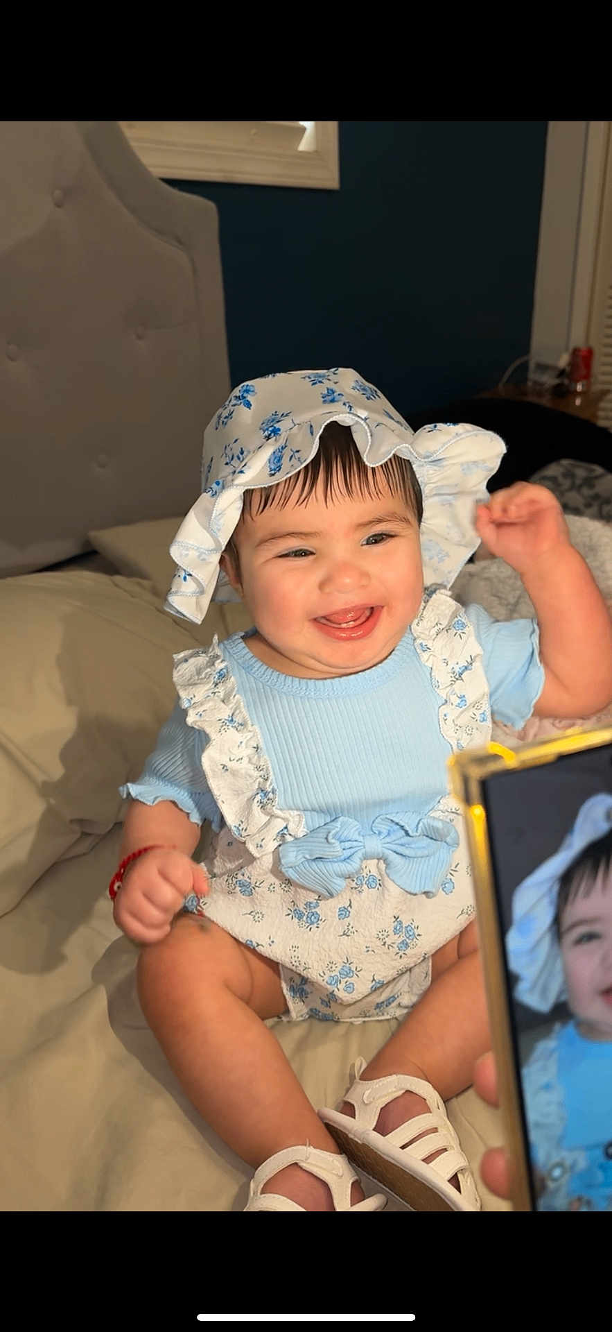 Aisha is registered to the contest to win money with this photo: baby, child, smiling, blue_clothing, bonnet, bed, indoor, person, happy, portrait, footwear, shoes, hand, skin, face, infant, cute, ruffles, flower_pattern, photography