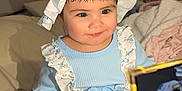 Aisha joined the competition — help win amazing prizes! baby, child, infant, smiling, blue_outfit, floral_pattern, bonnet, bed, blanket, phone, reflection, closeup, cute, indoors, person, sitting, white_sandals, bracelet, soft_lighting, portrait