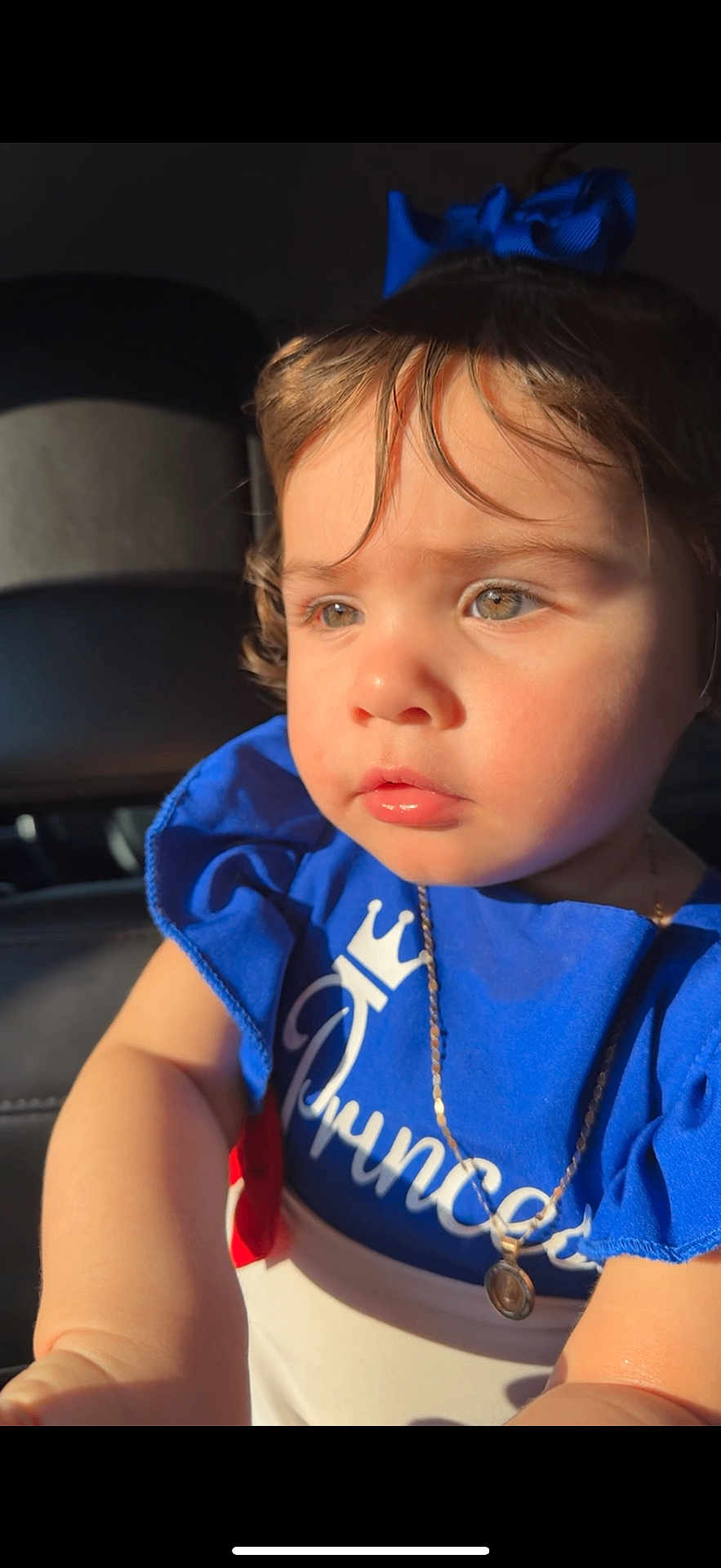Aisha joined the competition — help win amazing prizes! toddler, child, blue_clothing, bow, necklace, face, portrait, serious_expression, sunlight, car_interior, closeup, young_child, person, cute, fashion, accessory, hair, skin, eyes, sitting