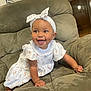 baby, child, smiling, happy, crawling, headband, bow, dress, white_clothing, floral_pattern, bracelet, armchair, green_furniture, indoor, home, cute, person, portrait, infant, young_child