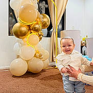 Benjamín is registered to the contest to win money with this photo: toddler, child, balloons, celebration, indoor, carpet, curtains, window, cross, dove, smile, shirt, jeans, shoes, adult_hands, decor, lighting, party, standing, happy