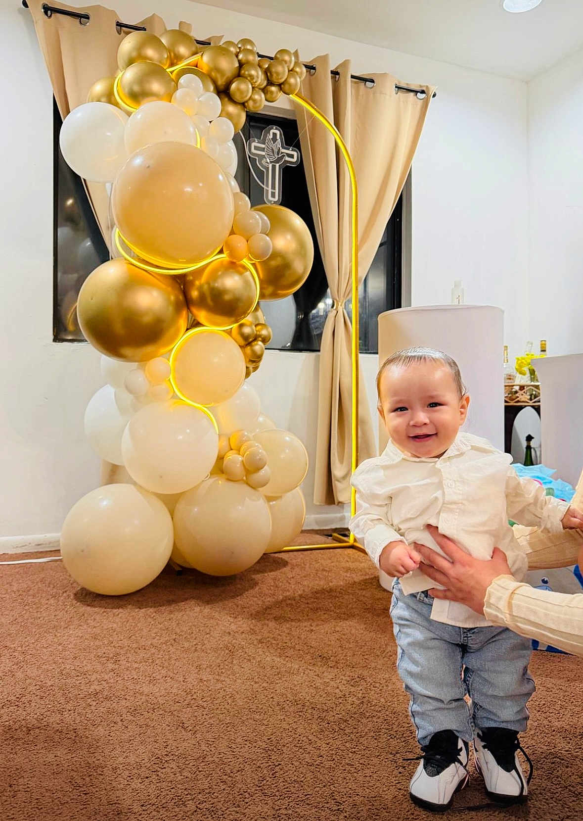 Benjamín is registered to the contest to win money with this photo: toddler, child, balloons, celebration, indoor, carpet, curtains, window, cross, dove, smile, shirt, jeans, shoes, adult_hands, decor, lighting, party, standing, happy