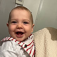 Liam a rejoint le concours — aidez-le/la à gagner de superbes lots ! baby, blanket, child, clothing, cute, face, hand, happy, holding, indoor, infant, light, person, portrait, skin, smiling, striped_overalls, teeth, wall, white_shirt