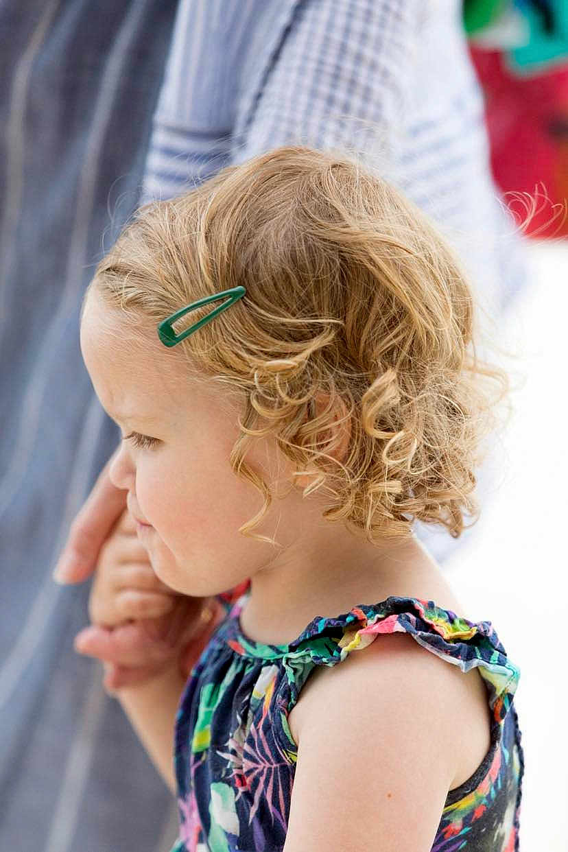 Issy is registered to the contest to win money with this photo: child, toddler, curly_hair, blonde, hair_clip, holding_hands, colorful_dress, floral_pattern, side_profile, portrait, outdoor, adult_hand, close_up, soft_background, cute, young_child, summer_clothing, skin, person, tender_moment