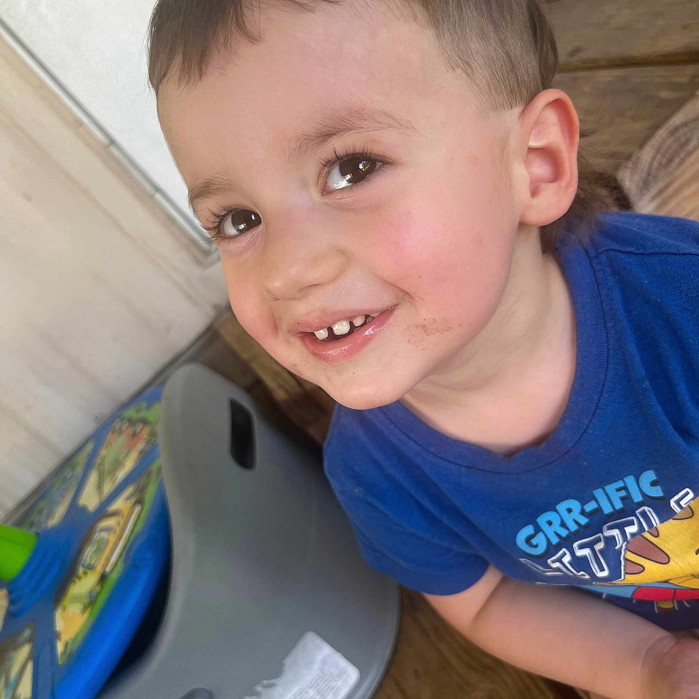 Rhyker is registered to the contest to win money with this photo: child, smiling, face, blue_shirt, messy_face, toy, chair, wooden_floor, indoor, happy, close_up, cute, young_child, casual_clothing, playful, portrait, person, toddler, brown_eyes, short_hair