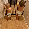 baby, baby_gate, back_view, barefoot, child, curly_hair, diaper, doorframe, hair, hallway, home, indoor, infant, light, room, safety_gate, standing, toddler, wall, wooden_floor