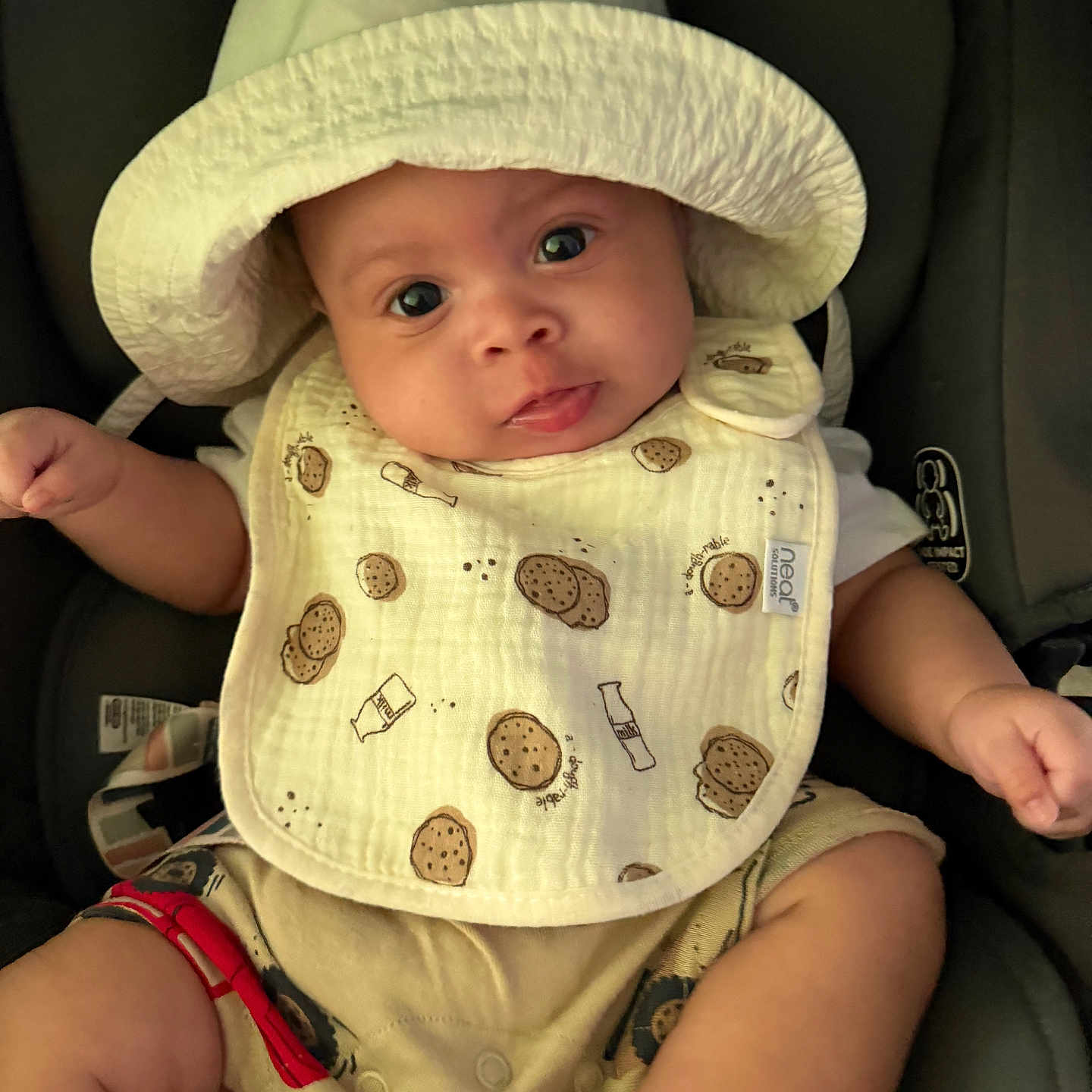Legend is registered to the contest to win money with this photo: baby, child, hat, bib, car_seat, cute, infant, clothing, sitting, face, eyes, hand, legs, footwear, white_hat, patterned_bib, closeup, indoors, adorable, portrait