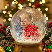 Jordan is registered to the contest to win money with this photo: child, snow_globe, christmas_tree, holiday, presents, red_hoodie, smiling, festive, decorations, pine_cones, lights, bokeh, winter, celebration, indoors, snowflakes, toy, gift_wrapping, ornaments, table