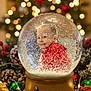 child, snow_globe, christmas_tree, holiday, presents, red_hoodie, smiling, festive, decorations, pine_cones, lights, bokeh, winter, celebration, indoors, snowflakes, toy, gift_wrapping, ornaments, table