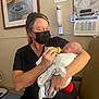 Jordan is registered to the contest to win money with this photo: woman, baby, feeding, bottle, hospital, mask, scrubs, newborn, medical_equipment, poster, chair, blanket, indoor, person, care, healthcare, nurse, holding, baby_wrap, tenderness