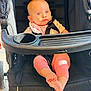baby, stroller, infant, child, pink_clothing, blue_eyes, feet, sitting, black_stroller, sunlight, curious_expression, seatbelt, outdoor, baby_bib, child_in_stroller, toddler, person, footwearless, daylight, baby_jogger