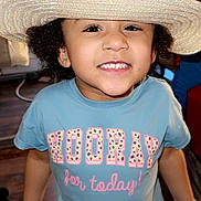 Aniyah is registered to the contest to win money with this photo: child, smiling, hat, straw_hat, indoor, tshirt, text_on_clothing, happy, person, face, curly_hair, brown_skin, young_child, portrait, casual_clothing, floor, window, furniture, home, bright_lighting