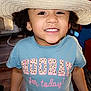 child, smiling, hat, straw_hat, indoor, tshirt, text_on_clothing, happy, person, face, curly_hair, brown_skin, young_child, portrait, casual_clothing, floor, window, furniture, home, bright_lighting