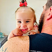 Rylee is registered to the contest to win money with this photo: baby, child, toddler, smile, face, hair_bow, red_bow, man, tattoo, shoulder, skin, freckles, tank_top, indoor, person, cute, playful, portrait, closeup, happy