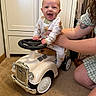 baby, child, toy_car, indoor, car, smile, person, pajamas, hand, carpet, furniture, cabinet, adult, supporting, happy, steering_wheel, vintage, playtime, cute, tattoo