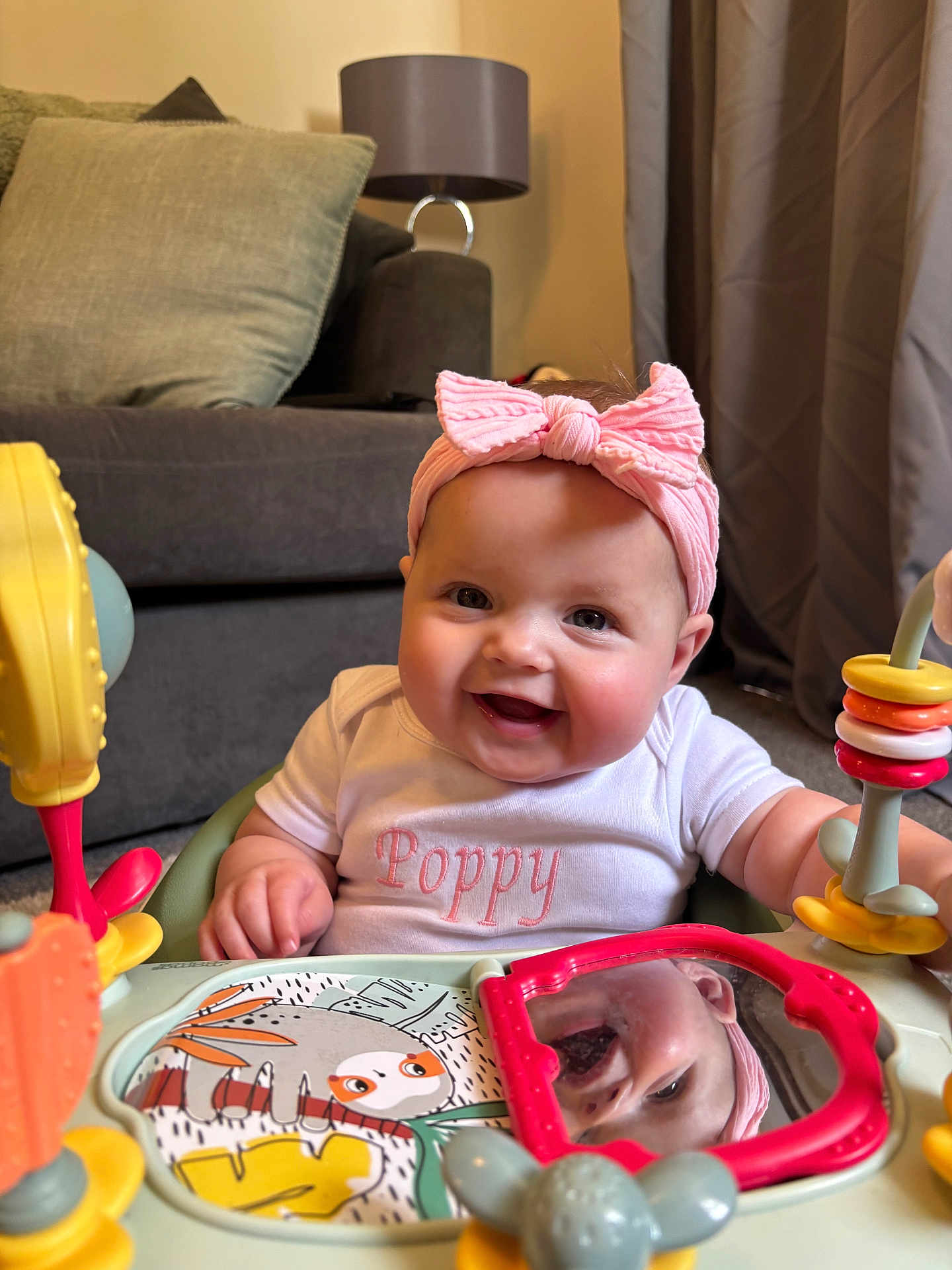 Poppy joined the competition — help win amazing prizes! baby, smiling, pink_headband, white_shirt, reflection, mirror, activity_center, toy, living_room, couch, cushion, lamp, curtains, child, infant, happy, indoors, person, play, cute