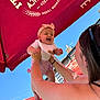 accessory, arms, baby, bib, blue_sky, brick_building, child, clothing, daytime, happy, headband, holding, joy, outdoor, person, playful, smiling, sunlight, umbrella, woman