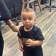 Jaxon is registered to the contest to win money with this photo: toddler, child, indoors, wooden_floor, black_tshirt, american_flag, camo_shorts, hairstyle, mohawk, hand, adult, supporting, curious, standing, casual_clothing, person, footwear, furniture, chair, floor