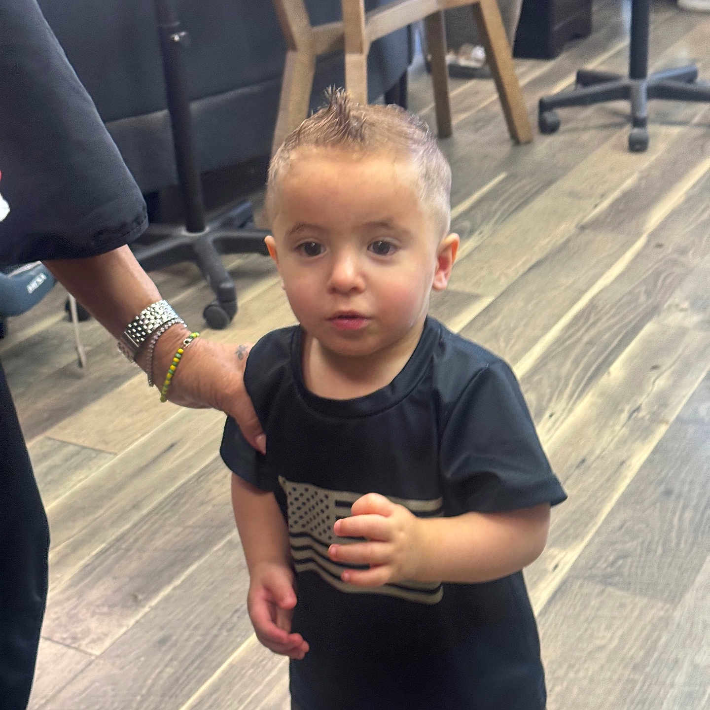 Jaxon is registered to the contest to win money with this photo: toddler, child, indoors, wooden_floor, black_tshirt, american_flag, camo_shorts, hairstyle, mohawk, hand, adult, supporting, curious, standing, casual_clothing, person, footwear, furniture, chair, floor