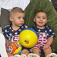 Jaxon is registered to the contest to win money with this photo: american_flag, ball, blue, chair, child, cushion, footwear, happy, indoor, matching_clothes, orange, person, playing, short_hair, sitting, smiling, stars, toddler, toy, yellow