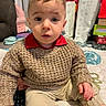 beige_pants, blanket, casual_clothing, child, curious, expression, floor, furniture, gifts, home, indoor, knitted_sweater, pattern, person, remote_control, seated, toddler, toys, window, young_child