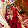 baby, infant, red_clothing, headband, blanket, christmas_tree, christmas_lights, plaid, floor, wood_floor, holiday, decorations, cute, indoors, person, newborn, festive, cozy, wrapped, relaxing