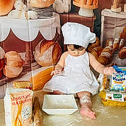 Sofia participe au concours pour gagner de l'argent avec cette photo : baby, child, chef_hat, white_dress, flour, rolling_pin, baking, kitchen, bread, pastries, eggs, milk_carton, table, food, cooking, messy, indoors, cute, person, apron