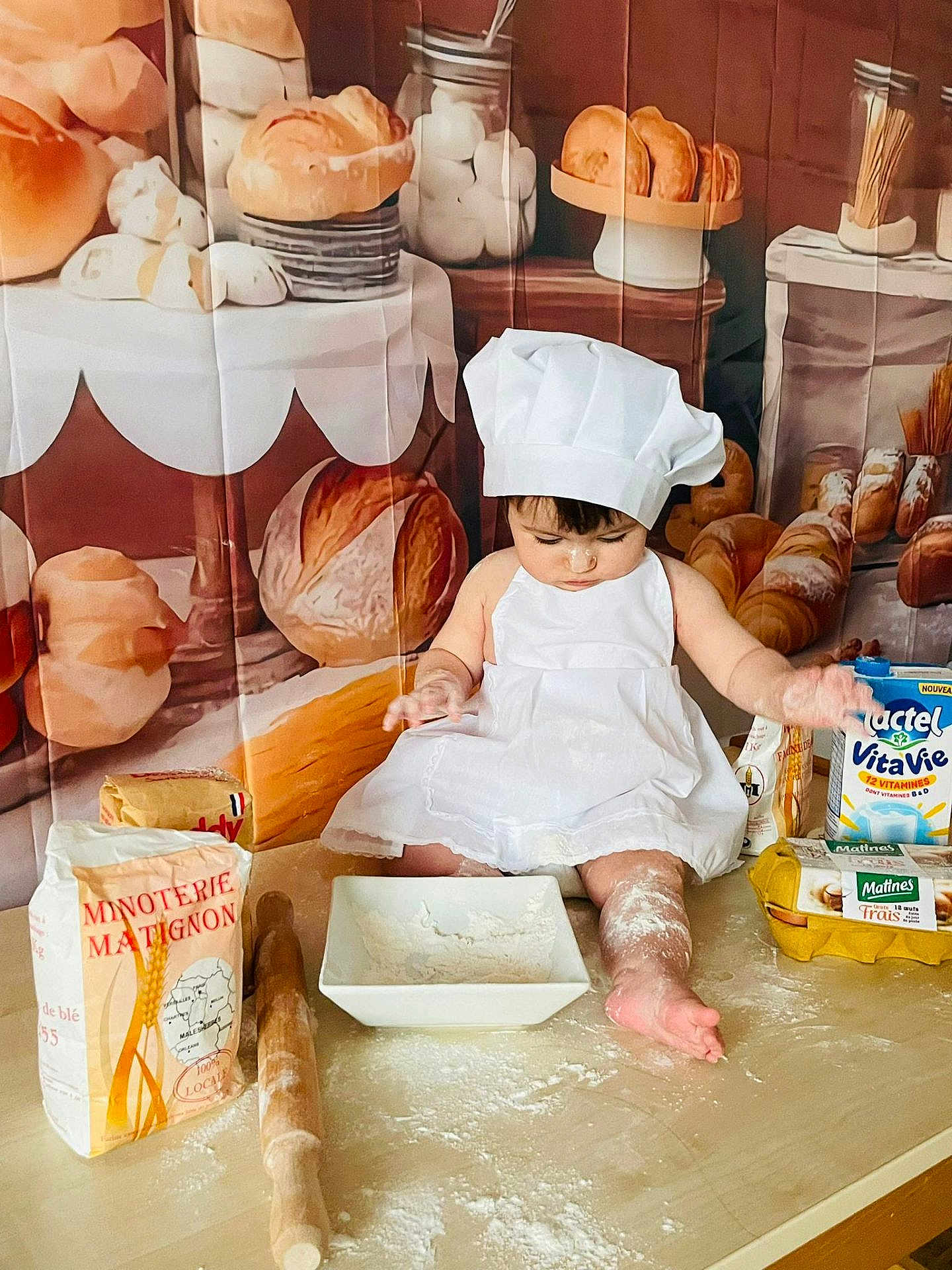 Sofia participe au concours pour gagner de l'argent avec cette photo : baby, child, chef_hat, white_dress, flour, rolling_pin, baking, kitchen, bread, pastries, eggs, milk_carton, table, food, cooking, messy, indoors, cute, person, apron