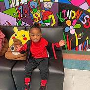 Jamar is registered to the contest to win money with this photo: child, smiling, sitting, black_couch, red_clothing, plush_toys, pikachu, poop_emoji_pillow, colorful_mural, wall_art, kindness_text, indoor, floor_tiles, happy, cute, casual_wear, sneakers, black_hair, playful, person