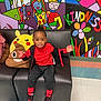 child, smiling, sitting, black_couch, red_clothing, plush_toys, pikachu, poop_emoji_pillow, colorful_mural, wall_art, kindness_text, indoor, floor_tiles, happy, cute, casual_wear, sneakers, black_hair, playful, person