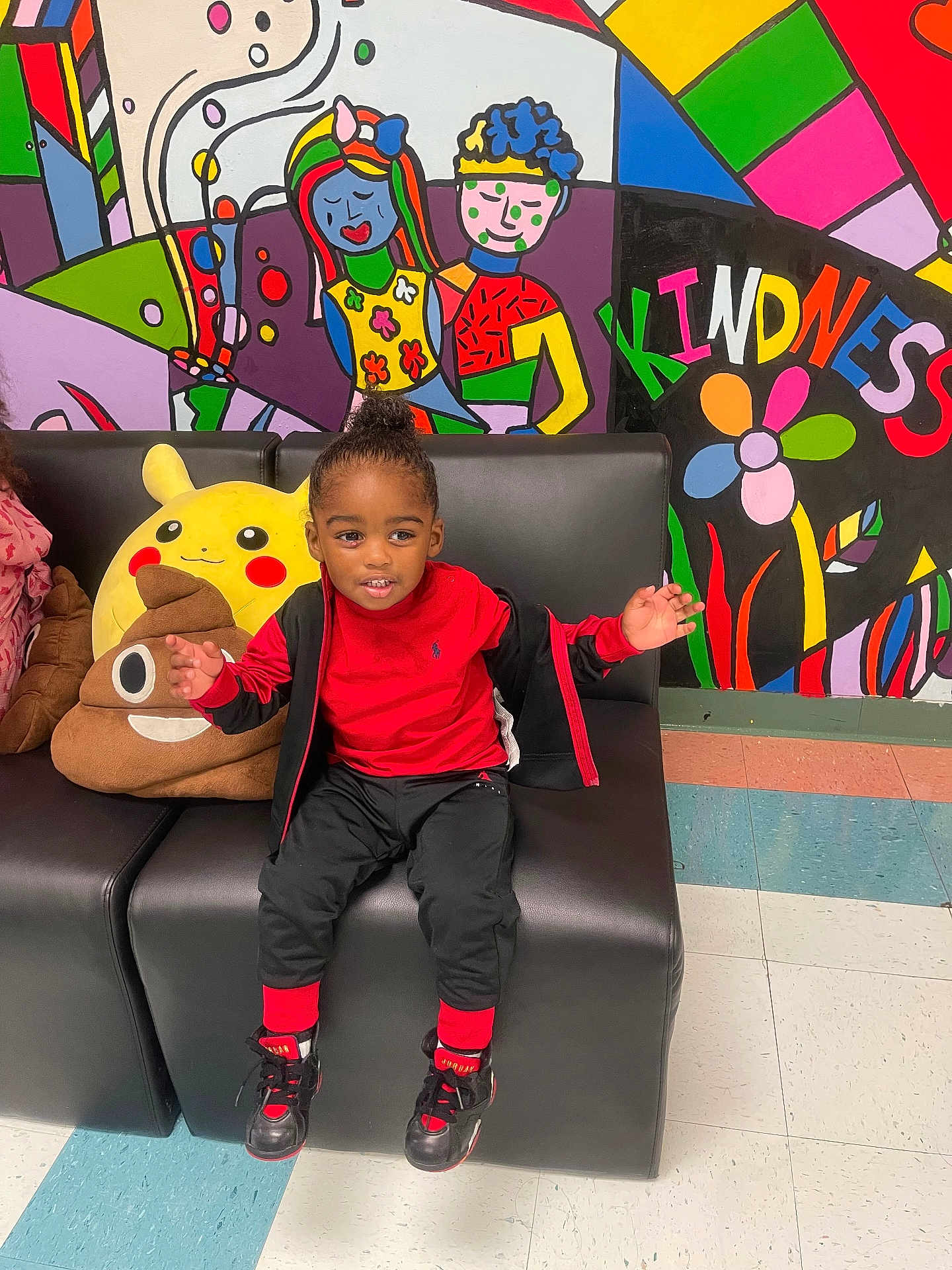 Jamar is registered to the contest to win money with this photo: child, smiling, sitting, black_couch, red_clothing, plush_toys, pikachu, poop_emoji_pillow, colorful_mural, wall_art, kindness_text, indoor, floor_tiles, happy, cute, casual_wear, sneakers, black_hair, playful, person