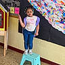 child, girl, stool, library, colorful_art, paper_feathers, peace_sign, playful, pouty_face, hair_beads, wall_art, bulletin_board, floor, jeans, sneakers, tshirt, indoor, school, hallway, expression