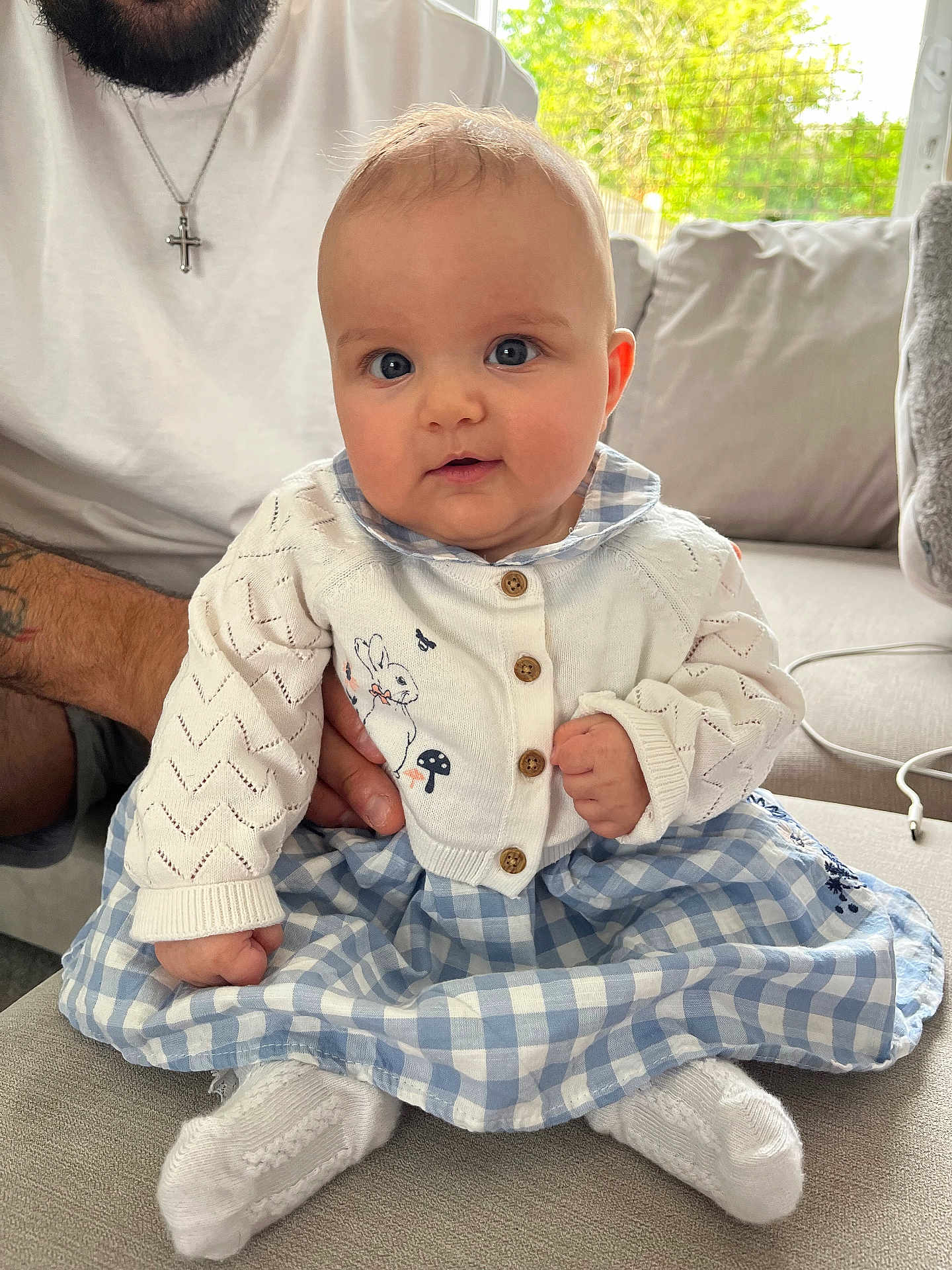Kiara is registered to the contest to win money with this photo: baby, child, blue_eyes, cardigan, dress, sitting, couch, indoor, hand, adult, white_socks, curly_hair, window, natural_light, tattoo, beard, cross_necklace, plaid, cute, portrait