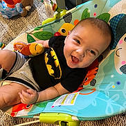 Oliver is registered to the contest to win money with this photo: baby, infant, child, smiling, happy, portrait, sitting, activity_seat, baby_toy, plush_toy, colorful, indoor, nursery, crib, carpet, clothing, black_shirt, gray_shorts, playtime, close_up