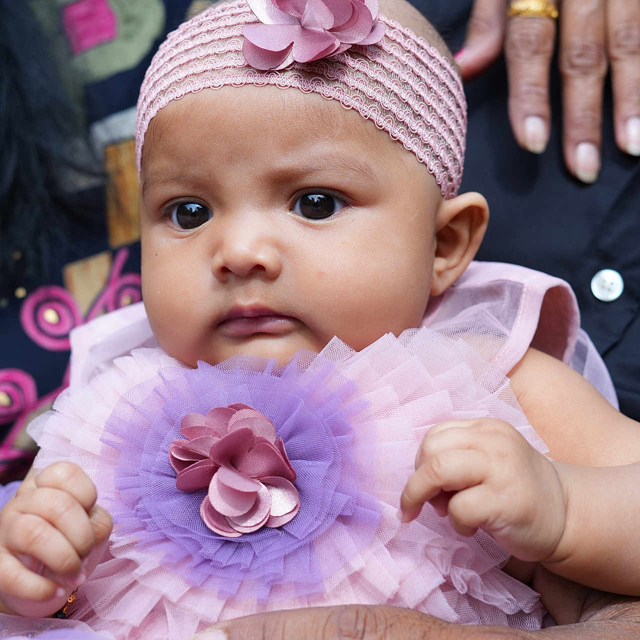 Jiyana is registered to the contest to win money with this photo: adult_hands, baby, child, closeup, colorful_background, cute, dress, expression, fabric, face, flower_accessory, hands, infant, outdoor, person, pink_headband, portrait, purple_tulle, skin, soft_light