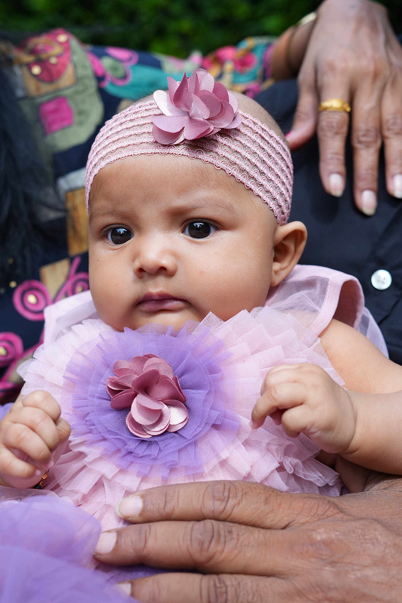 Jiyana is registered to the contest to win money with this photo: baby, child, infant, pink_headband, flower_accessory, purple_tulle, hands, adult_hands, dress, closeup, portrait, outdoor, face, cute, person, skin, expression, soft_light, colorful_background, fabric