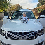 Julius Madison is registered to the contest to win money with this photo: baby, child, car, range_rover, hood, clothing, beanie, smiling, outdoor, vehicle, door, white_car, automobile, tree, sky, daylight, happy, person, sitting, side_mirror