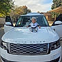 baby, child, car, range_rover, hood, clothing, beanie, smiling, outdoor, vehicle, door, white_car, automobile, tree, sky, daylight, happy, person, sitting, side_mirror