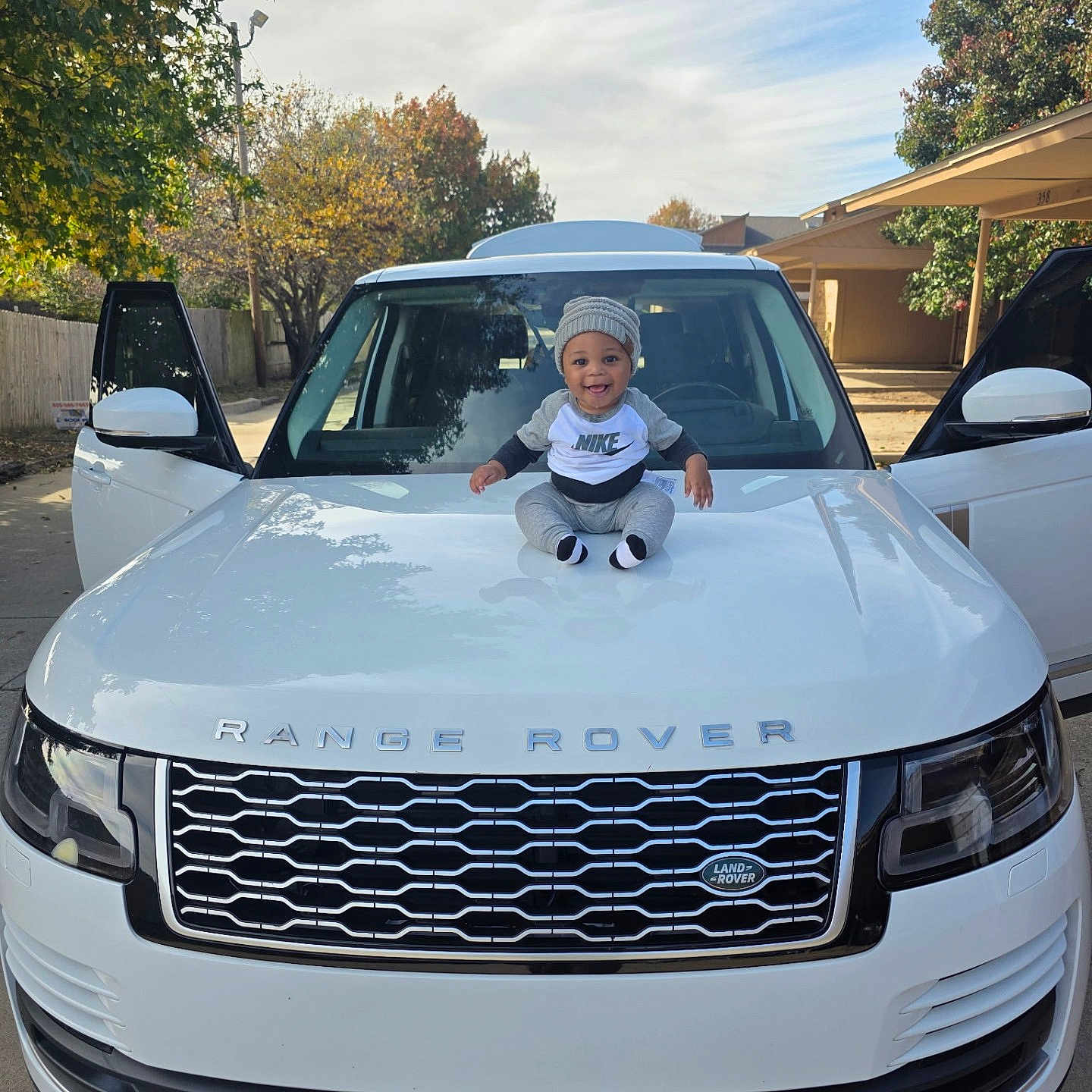 Julius Madison is registered to the contest to win money with this photo: automobile, baby, beanie, car, child, clothing, daylight, door, happy, hood, outdoor, person, range_rover, side_mirror, sitting, sky, smiling, tree, vehicle, white_car