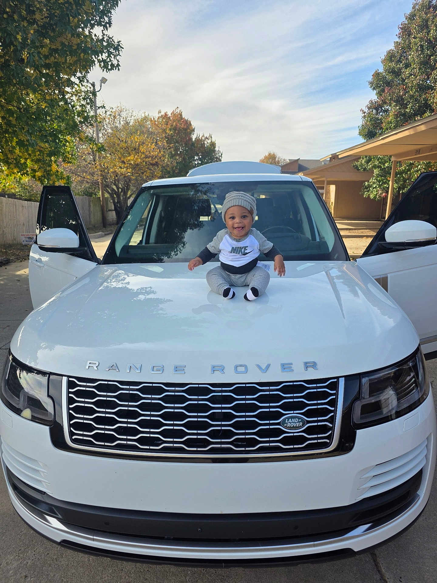 Julius Madison is registered to the contest to win money with this photo: baby, child, car, range_rover, hood, clothing, beanie, smiling, outdoor, vehicle, door, white_car, automobile, tree, sky, daylight, happy, person, sitting, side_mirror
