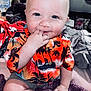 baby, child, infant, smiling, sitting, hand_in_mouth, orange_shirt, hawaiian_shirt, patterned_clothing, shorts, indoor, blanket, bed, cute, happy, portrait, face, blue_eyes, light_skin, playful